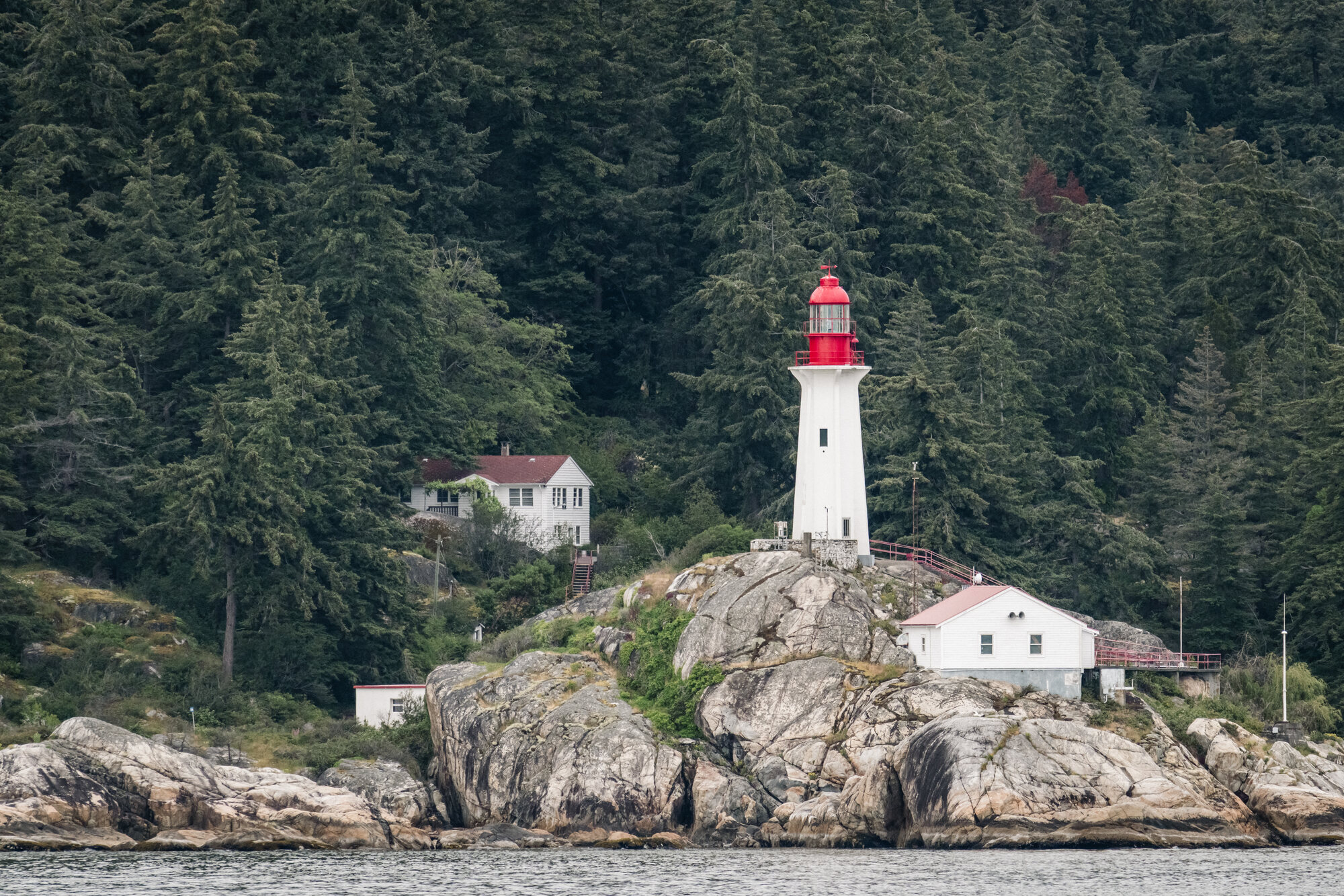White Point Atkinson Lighthouse with a red lantern standing on granite below dense coastal forest