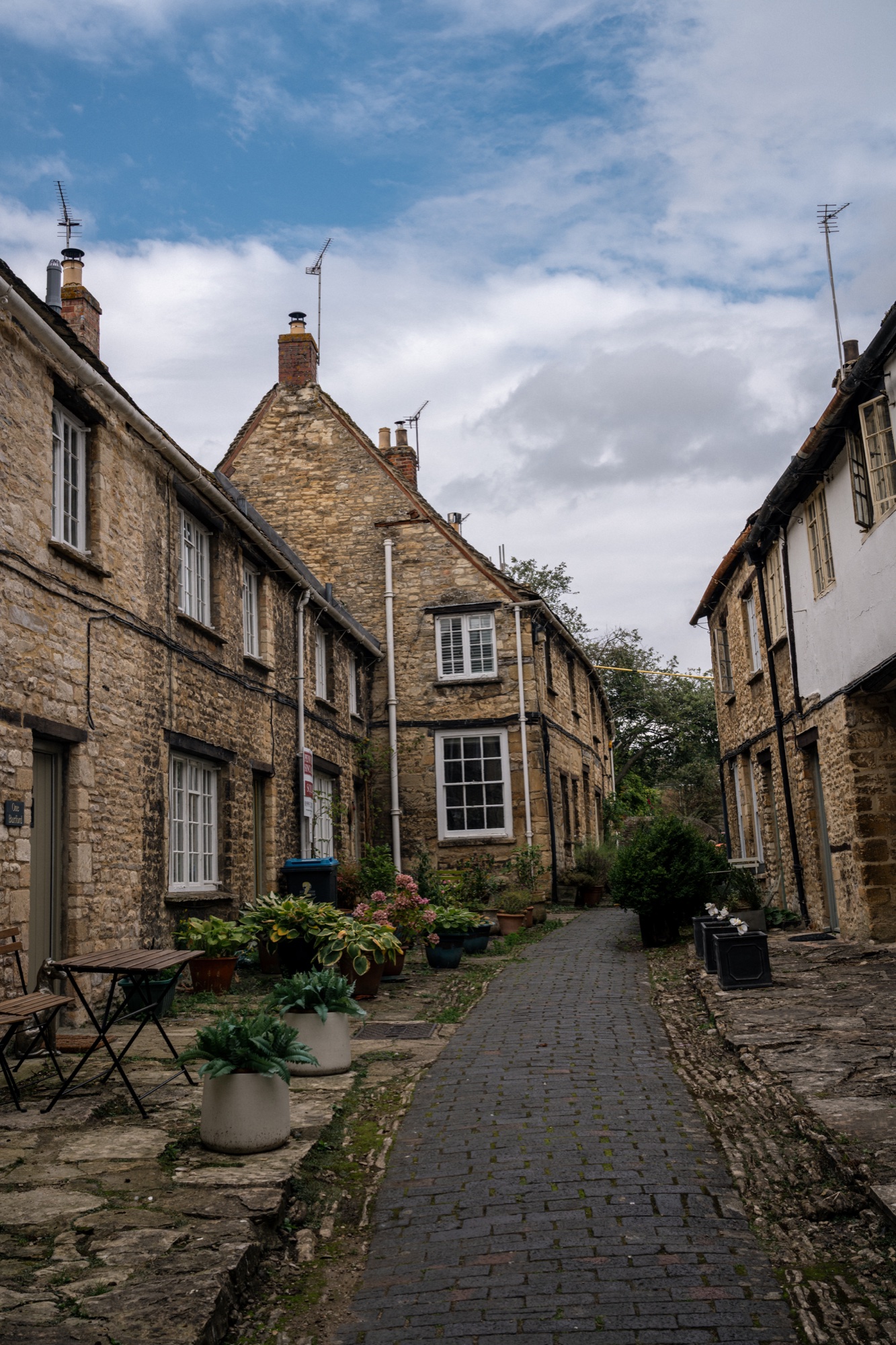 Narrow pedestrian lane between Cotswold stone cottages with flowers and a blue sky overhead