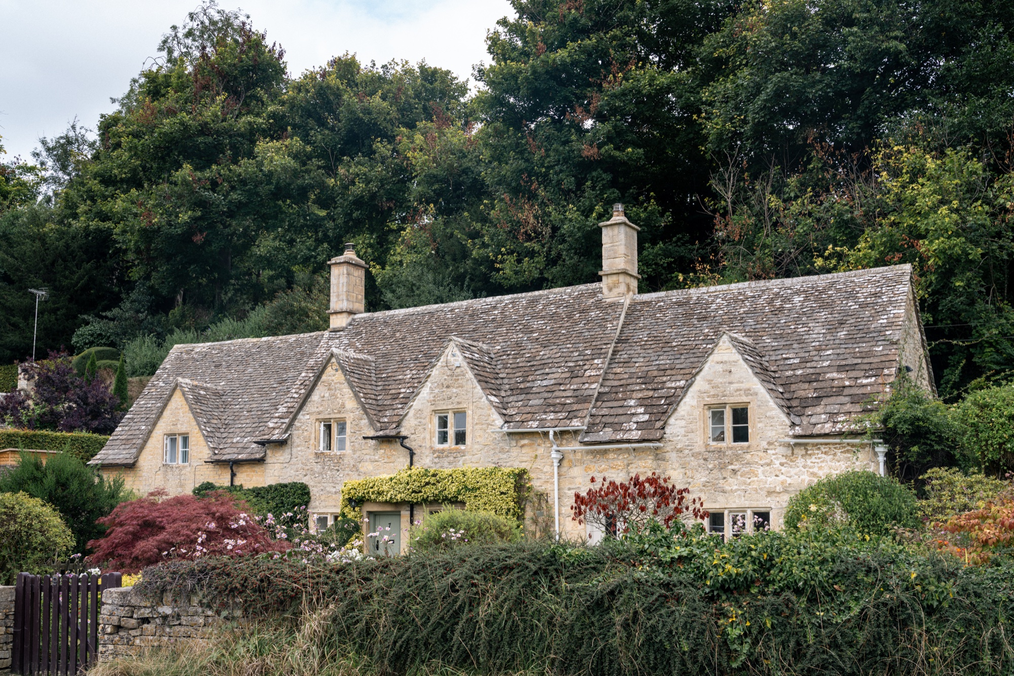 Honey-colored Cotswold stone cottages surrounded by layered trees and a colorful garden