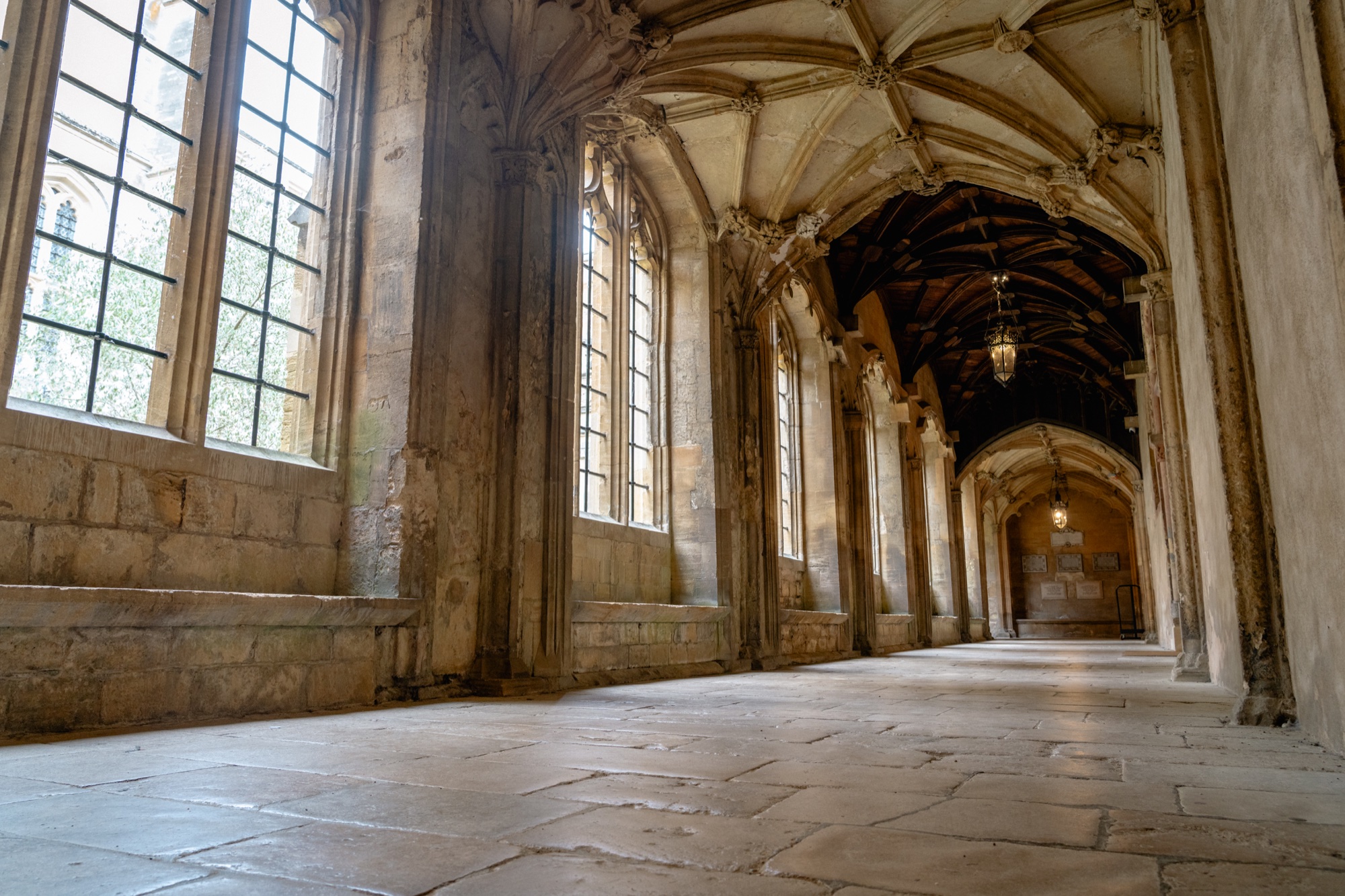 Sunlit Oxford stone cloister with Gothic arches, tall windows, and a worn flagstone floor